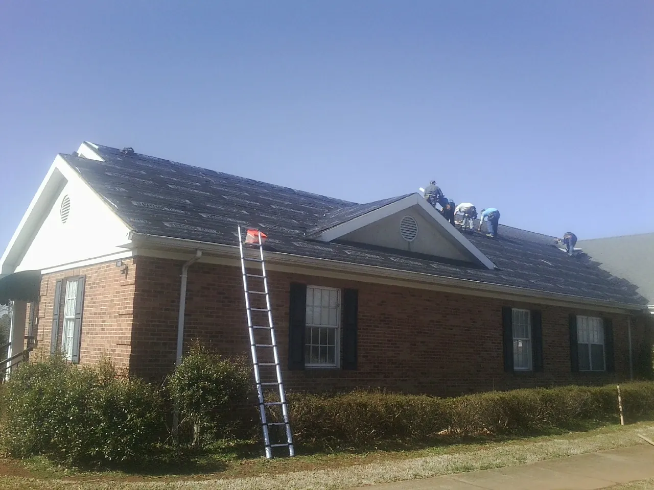 Crew tearing shingles off a brick ranch roof with a ladder set — Emergency Roof Repair work by our crew in Elbridge