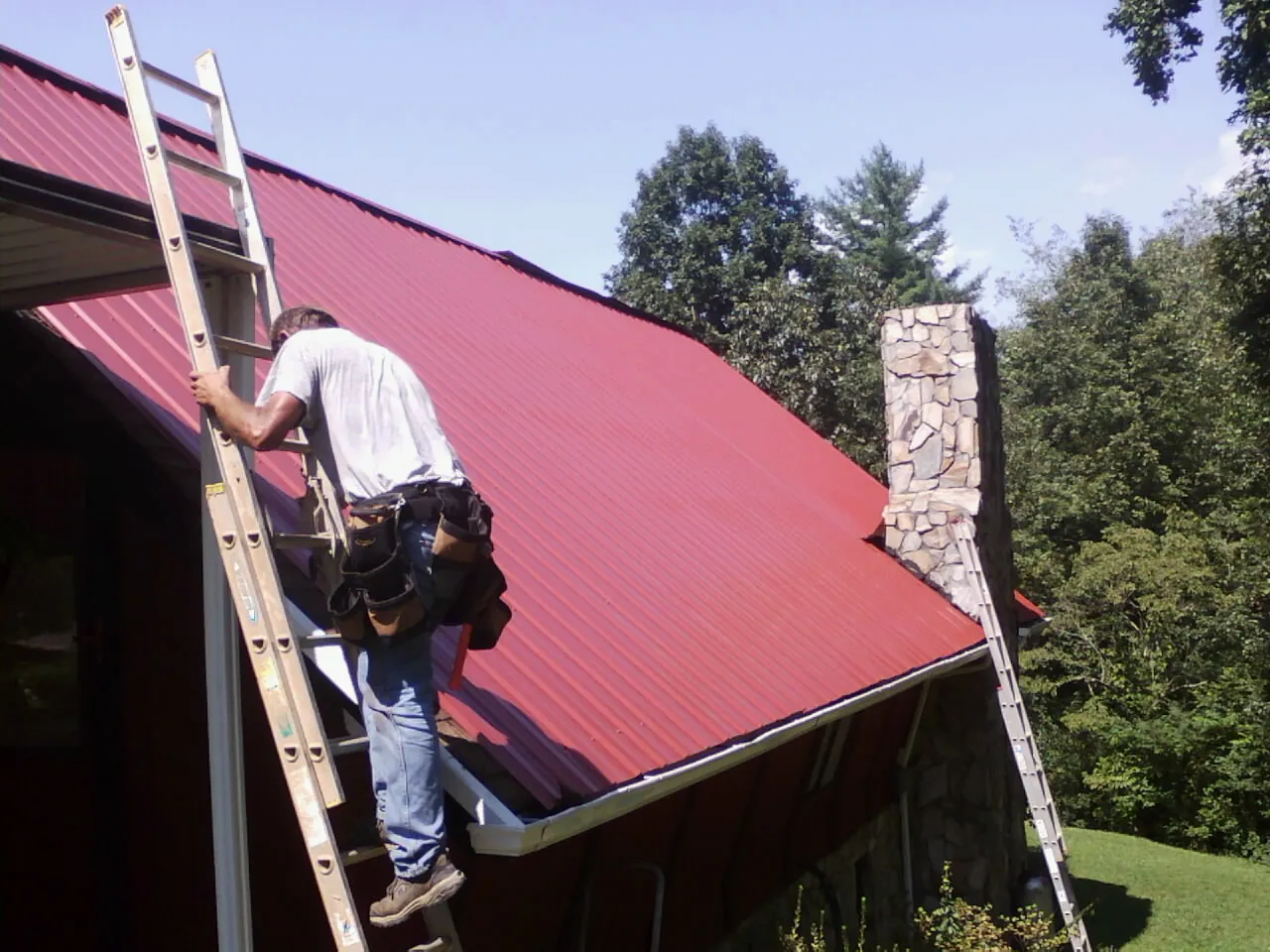 Roofer on a ladder beside a red metal roof and stone chimney — Soffit & Fascia Repair by Roofing Brothers in Elbridge
